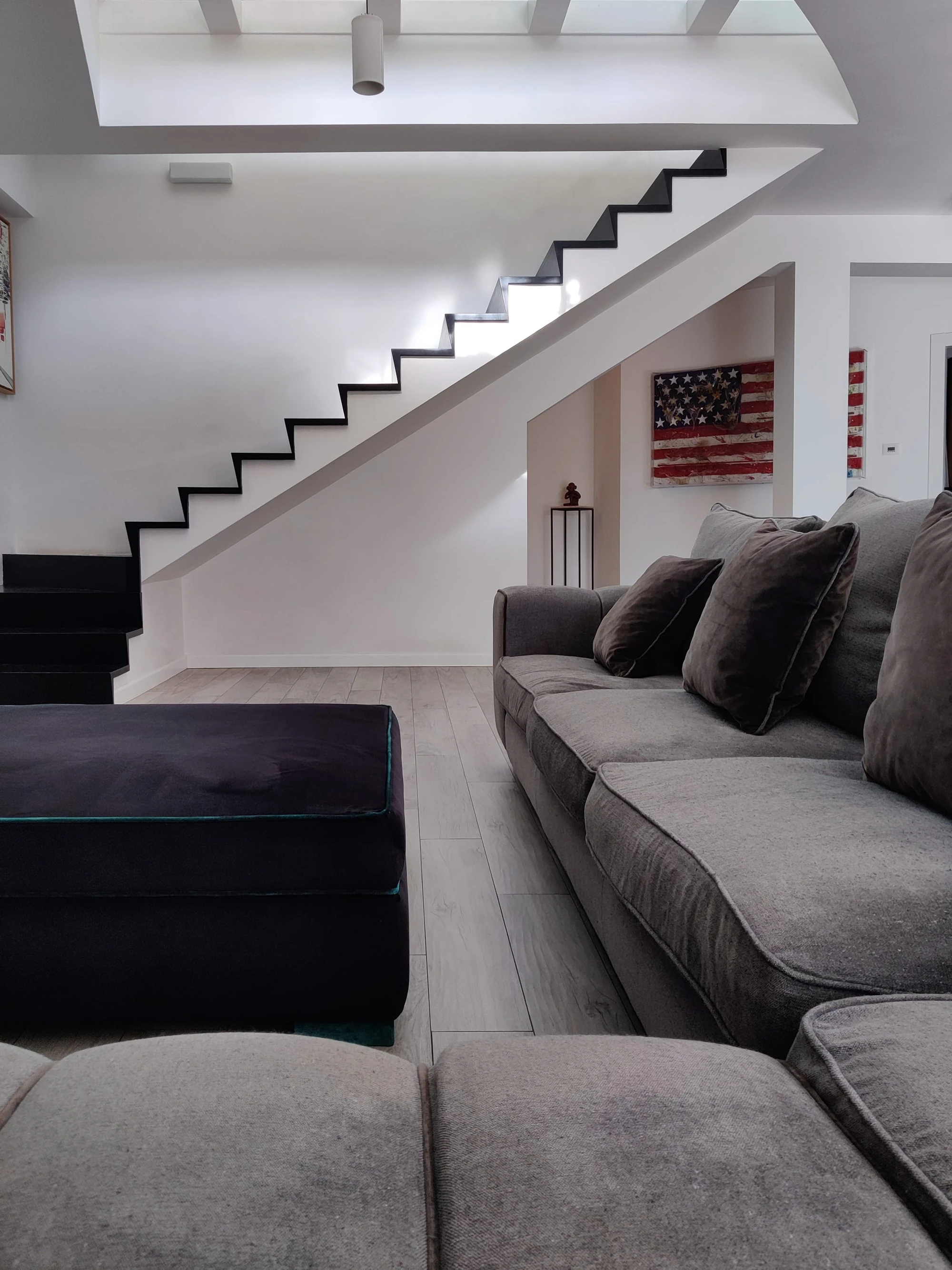 Soggiorno open space DS Villa: divano e pouf in velluto grigio scuro, scala a sbalzo in metallo nero su parete bianca, parete decorativa con bandiera americana vintage e pavimento in legno chiaro