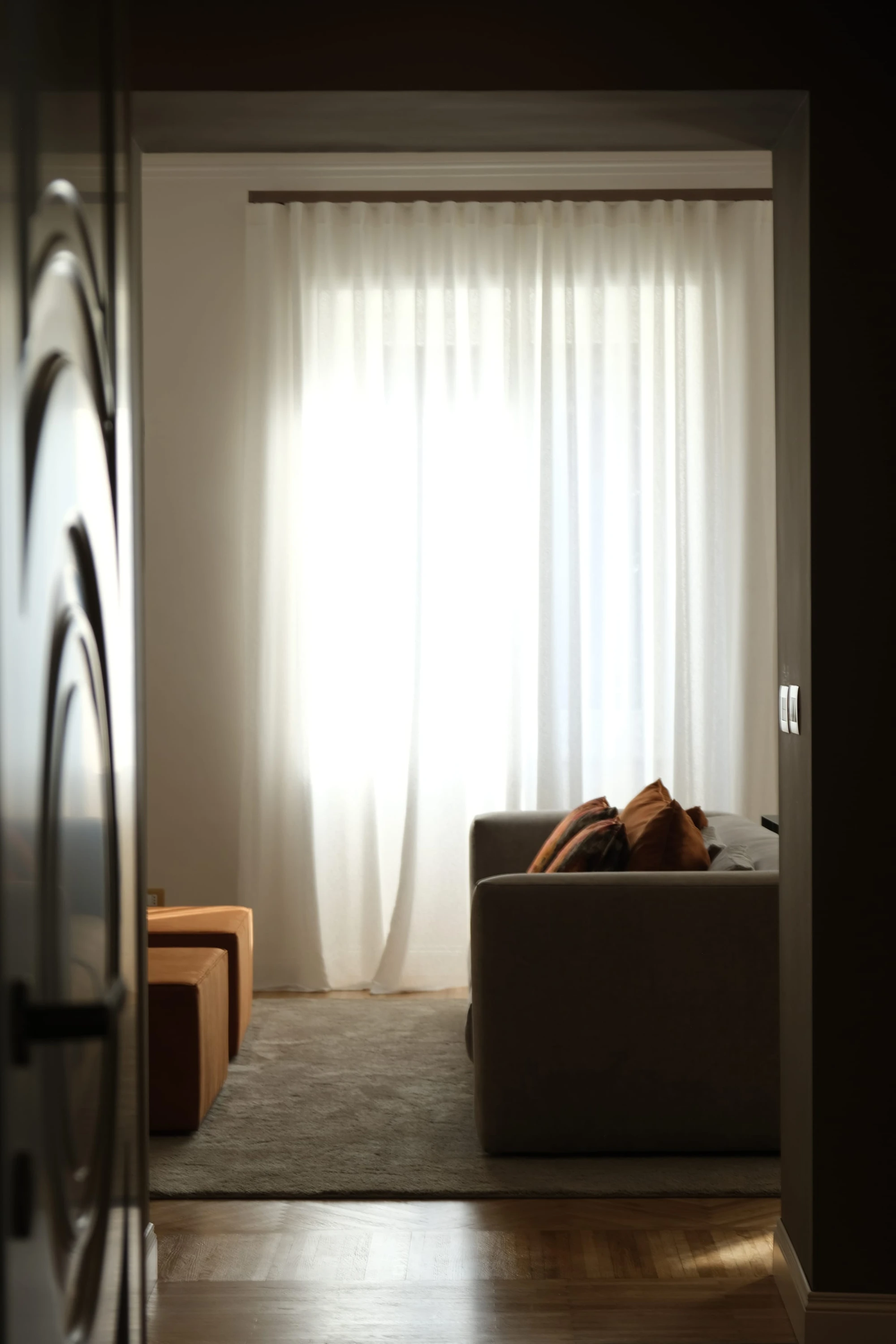 Casa vacanze SC: vista verso il soggiorno attraverso una soglia in penombra, tende bianche filtranti che diffondono luce calda su divano grigio con cuscini decorativi e parquet a spina di pesce